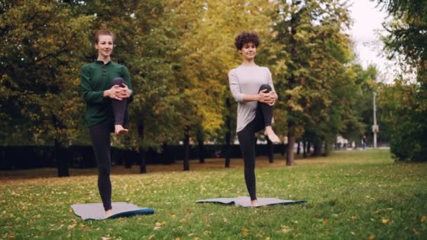 Damas delgadas están haciendo ejercicio al aire libre en el parque de pie en una pierna practicando Padangusthasana durante la clase individual con un profesor experimentado. Naturaleza y concepto de juventud . — Vídeos de Stock