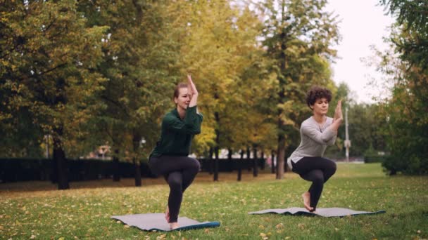 Instructora de yoga femenina está enseñando a alumna principiante a posar águila en el parque, chica alegre está cometiendo errores y riéndose, las mujeres están disfrutando de la naturaleza y el ejercicio . — Vídeos de Stock