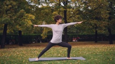 Kısa kıvırcık saçlı güzel kız açık havada savaşçı poz ayakta sonra park pratikte sırasında vücut ve bacak Germe yoga yapıyor. Ağaç ve otların görülebilir.