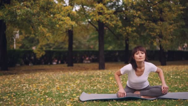 La joven flexible está haciendo ejercicios para la columna vertebral saludable sentada en la esterilla de yoga y moviéndose hacia atrás y el cuerpo durante la práctica individual en el parque. Concepto de salud y naturaleza . — Vídeo de stock