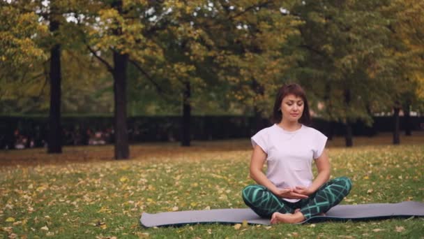 Ganska ung kvinna är avkopplande sitter i lotus pose på yogamattan i park och andas vila efter enskilda praktiken. Meditation och naturen koncept. — Stockvideo