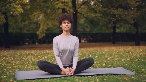 La guapa morena está estirando las piernas en zapatillas posando sentada en una esterilla de yoga y luego inclinándose hacia adelante relajándose y cerrando los ojos. Concepto de relajación y yoga . — Vídeo de stock