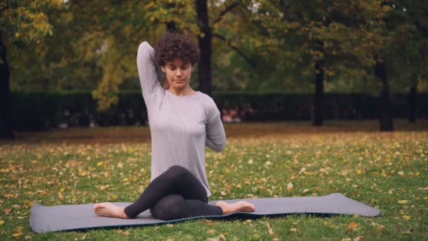 Eine ernsthafte junge Yogini trainiert im Park sitzend in Kuhgesicht-Pose mit den Armen auf dem Rücken beim Freiluft-Training im Stadtpark. Natur- und Gesundheitskonzept. — Stockvideo