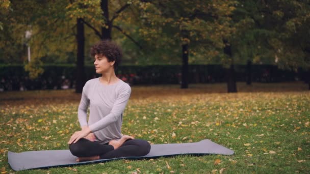 Ładna dziewczyna w Sport Odzież jest skręcanie jej ciało, siedząc w pozycji lotosu, a następnie relaks z zamkniętymi oczami po praktyce w parku na jesienny dzień. — Wideo stockowe