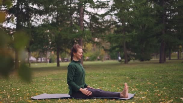 Glimlachend jonge vrouw is op park doen yoga zittend op de mat op groene en gele gras genieten van frisse lucht en gezonde activiteit uitoefenen. Jeugd en recreatie concept. — Stockvideo