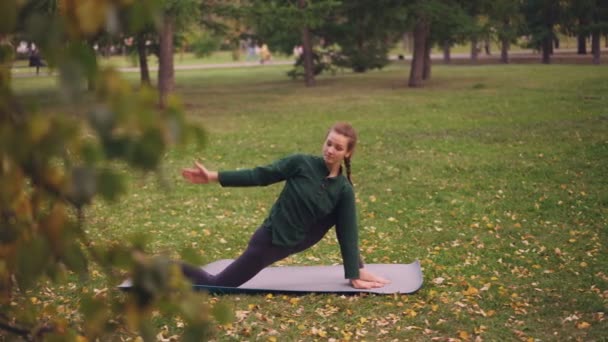 Mujer bastante joven se concentra en la práctica de yoga estirando las piernas y el equilibrio en la formación de esterillas de yoga solo en el parque en otoño. Concepto de relajación y salud . — Vídeos de Stock
