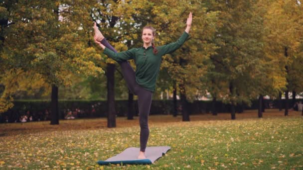 Lachende meisje professionele yoga-instructeur doet evenwicht oefeningen staan op één been op mat op gras in park. Mooie herfst natuur is zichtbaar. — Stockvideo