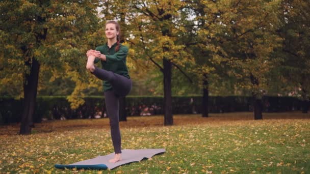 Estudiante de yoga femenino está balanceándose en una pierna de pie en la estera en el área recreativa haciendo ejercicio solo en el día de otoño. Concepto moderno de juventud, salud y naturaleza . — Vídeo de stock