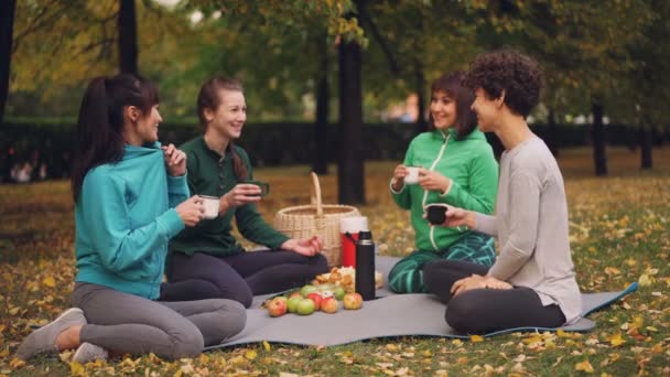 Glada flickor har picknick i parken sitter på yogamattor och äta efter utomhus praxis under hösten, flickor talar och skrattar. Kommunikation och mat koncept. — Stockvideo