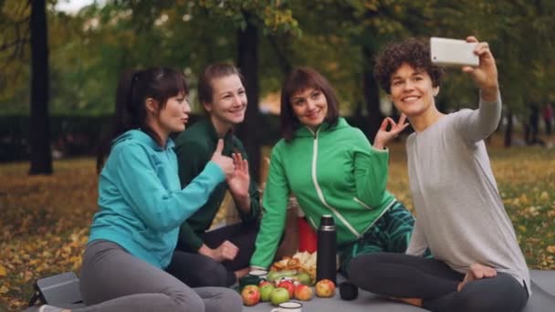 Jovens atraentes yogini estão tomando selfie usando smartphone durante piquenique no parque no outono. Meninas estão posando e sorrindo se divertindo sentado em tapetes . — Vídeo de Stock