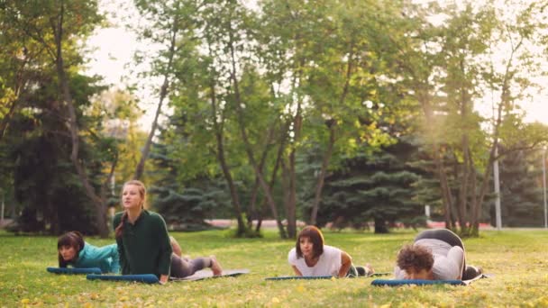 Slanka flickor i sportkläder gör yoga tillsammans i grupp utomhus klass i park utvecklar styrka och flexibilitet. Millenniegenerationen och aktiv livsstilskoncept. — Stockvideo