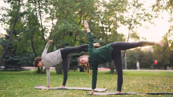 Piękne młode panie stoją w Half Moon stanowią Ardha Chandrasana trakcie parą zajęcia jogi w parku. Zdrowy styl życia, nauczania i koncepcja nauki i sportu. — Wideo stockowe