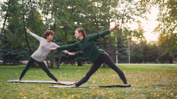 Instructora experimentada chica delgada está enseñando a su estudiante hatha yoga asanas en el cálido día de otoño en el parque de la ciudad. Estilo de vida saludable y deportes para la gente urbana concepto . — Vídeo de stock