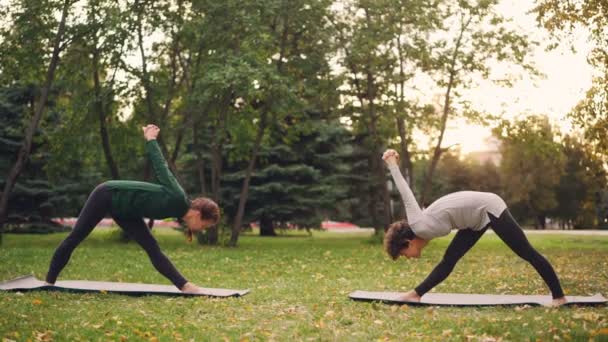 Yoga student is exercising outdoors with instructor stretching legs and back bending forward standing on mats on green and yellow meadow in city park. — Stock Video