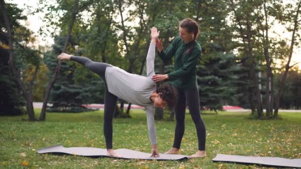 Freundliche Yogalehrerin hilft Studentin, Halbmond-Pose beizubehalten ardha chandrasana steht neben ihr, spricht und hält ihre Hand und ihr Bein. — Stockvideo