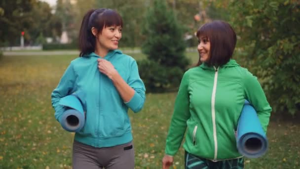 Dolly toma de deportistas guapos hablando, sonriendo y sosteniendo esterillas de yoga caminando en el parque en el día de otoño. Gente urbana, ocio y deporte . — Vídeos de Stock