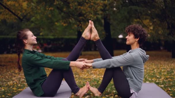 Attractive girls are doing yoga exercises in pair holding hands and putting feet together raising legs sitting on mat in park. Enjoyable training and people concept. — Stock Video