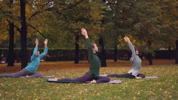 Groep van jonge dames doet yoga praktiserende koning Pigeon pose Eka Pada Rajakapotasana op matten op mooie groen en gele gazon in een park op herfstdag. — Stockvideo
