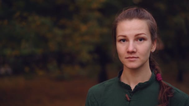 Ritratto ravvicinato di bella ragazza con treccia che guarda la macchina fotografica e sorride poi ridendo in piedi all'aperto nel parco in autunno con alberi sullo sfondo . — Video Stock