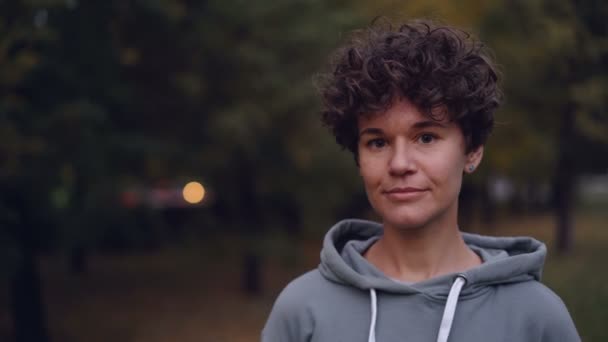 Portret van knap meisje met kort krullend haar kijken camera lacht dan lachen staande buiten in herfst in stadspark. Mens en natuur concept. — Stockvideo