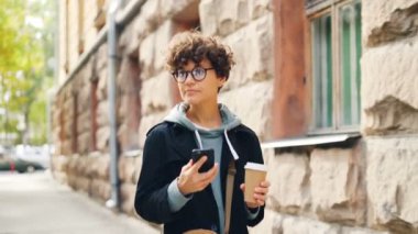 Siyah kıvırcık saçlı güzel kız smartphone manifatura sokulgan istimal ve kahve şehrinde yürüyüş tutarak. Modern teknoloji, iletişim ve içecekler kavramı.