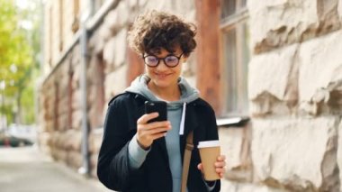 Son moda gözlük gülümseyen genç kadın açık havada gitmek kahve ile büyük şehirde yürürken ekrana bakarak akıllı telefon kullanıyor. Gençlik yaşam tarzı, street ve teknoloji kavramı.