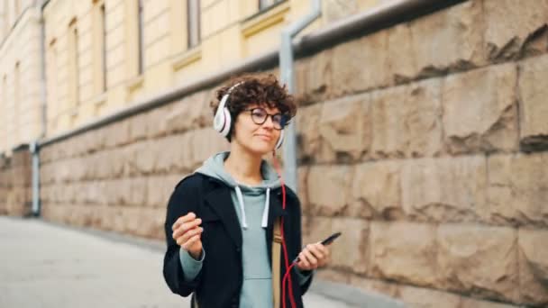 Όμορφος μελαχρινή σε κομψά ποτήρια είναι Ακρόαση ραδιοφώνου στα ακουστικά και χρησιμοποιώντας το smartphone κατά τη διάρκεια της βόλτα στην πόλη οικογένεια το φθινόπωρο. Άνθρωποι και έννοια αστική ζωή. — Αρχείο Βίντεο