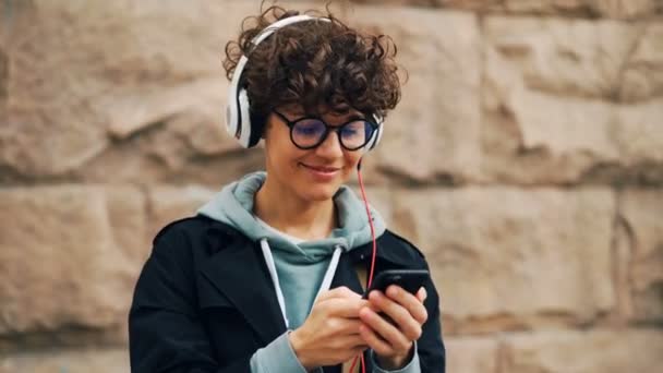 Menina bonita é fones de ouvido e óculos está ouvindo música e tocando a tela do smartphone escolhendo músicas de pé ao ar livre na cidade com parede de pedra no fundo . — Vídeo de Stock