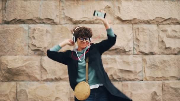 Feliz joven melómana está cantando y bailando escuchando música en auriculares y sosteniendo el teléfono inteligente al aire libre en la calle. Canciones y concepto juvenil . — Vídeos de Stock