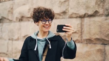 Neşeli kız video seslenmek, konuşma ve holding smartphone mimik ve kulaklık giyen ekrana bakarak yapıyor. Modern teknoloji, iletişim ve insan kavramı.