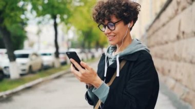 Kıvırcık saçlı ve gözlüklü genç kadınla online açık havada duran ve akıllı telefon ve kulaklık kullanarak arkadaşlarıma konuşuyor. Becerikli, internet ve iletişim kavramı.