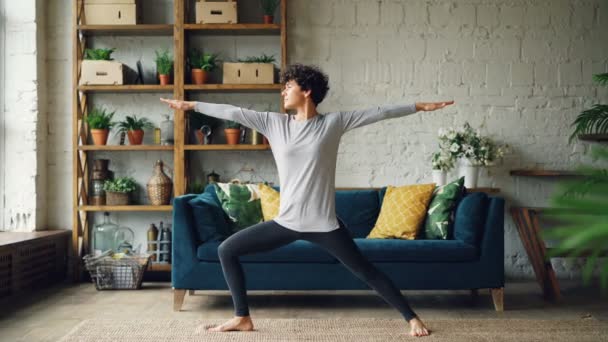 Стройная молодая женщина практикует йогу дома, стоя в позиции Воина, развивая силу и выносливость. Квартира в мансардном стиле с мебелью видна . — стоковое видео