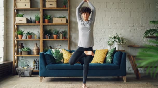 Bella ragazza è concentrata sulla pratica dello yoga a casa facendo esercizi di bilanciamento in piedi sul pavimento su una gamba. Stile di vita sano, concetto di gioventù e appartamento . — Video Stock