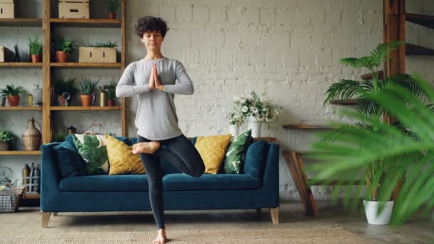 Die attraktive junge Frau balanciert allein beim Yoga zu Hause auf einem Bein in Baumstellung mit den Händen in der Hand. Gesunde Menschen, Wohnungen und Sportkonzept. — Stockvideo