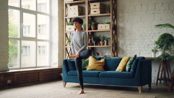 Slim yogini mulher está fazendo ioga sozinho no apartamento estilo loft moderno equilibrando em uma perna com as mãos levantadas em Namaste. Belo interior é visível . — Vídeo de Stock