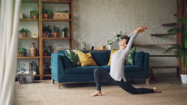 Jonge dame in sportkleding doet lichaamsoefeningen stretching benen en lichaam uitoefenen op verdieping in het mooie appartement. Sportieve concept van jeugd en gezondheid. — Stockvideo
