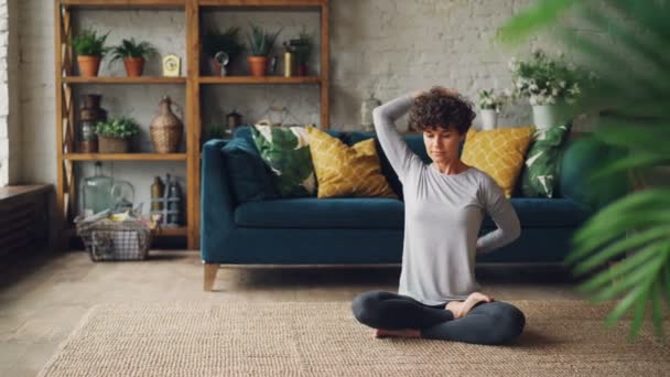 Bonita senhora está esticando os braços atrás das costas sentada no chão em posição de lótus durante o treinamento pessoal em casa. Estilo de vida ativo, pessoas e conceito de saúde . — Vídeo de Stock