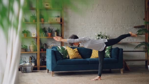 Mujer joven delgada se concentra en ejercicios de equilibrio durante el entrenamiento personal en casa de pie en el suelo en una pierna. Bienestar y concepto deportivo . — Vídeos de Stock