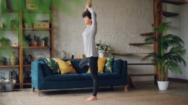 Esnek ve güçlü genç kadın yükseltilmiş silah ile denge kurmak tek ayak üzerinde duran yoga yapıyor evde eğitiyor. Sağlıklı yaşam tarzı ve iç kavramı.