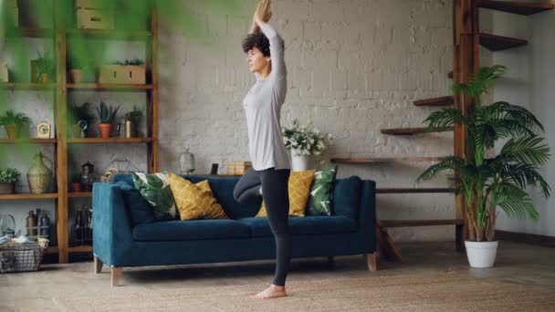 Mujer joven flexible y fuerte está entrenando en casa haciendo yoga de pie sobre una pierna manteniendo el equilibrio con los brazos levantados. Concepto de estilo de vida saludable e interiores . — Vídeos de Stock