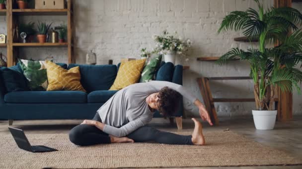 Красива дівчина дивиться підручники йоги на екрані ноутбука і займається фізичними вправами, сидячи на підлозі вдома наодинці. Сучасні технології, саморозвиток та молодіжна концепція . — стокове відео