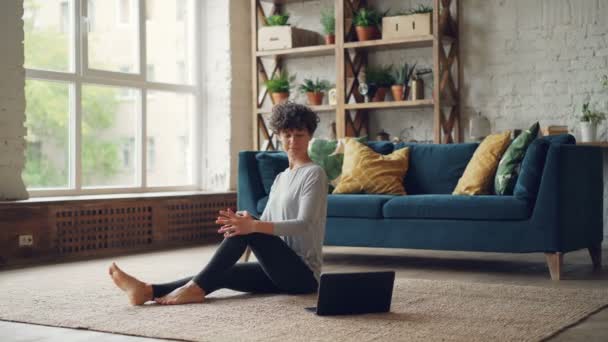 La joven bonita está aprendiendo yoga de lecciones en línea sentada en el piso mirando la pantalla del ordenador portátil y practicando giros corporales. Concepto de Internet, deportes y hobby . — Vídeos de Stock