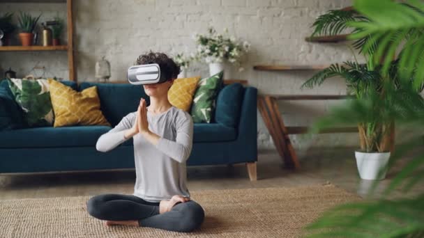 Menina esbelta em óculos de realidade virtual está meditando sentado no chão em casa em posição de lótus e desfrutando de prática pessoal. Tecnologia moderna e conceito de ioga . — Vídeo de Stock