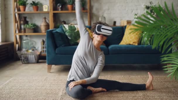 Lächelndes Mädchen mit Virtual-Reality-Brille sitzt zu Hause auf dem Boden und streckt Beine und Körper und genießt es. gesunder Lebensstil und vr-Simulationskonzept. — Stockvideo