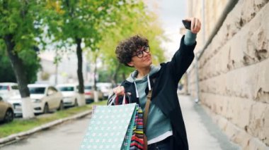 Sokakta duran, akıllı telefon tutarak ve kamera için poz gülümseyen poşetler ile selfie alarak güzel kız ağır çekim. Şehir ve teknoloji kavramı.