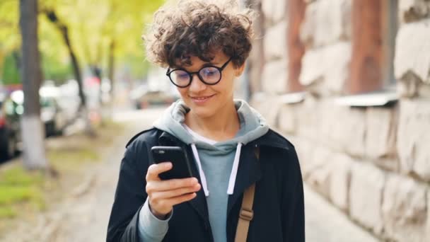 Movimento lento de mulher sorridente em óculos e roupas da moda usando smartphone olhando para a tela e andando ao ar livre na cidade. Estilo de vida juvenil, conceito de rua e tecnologia . — Vídeo de Stock