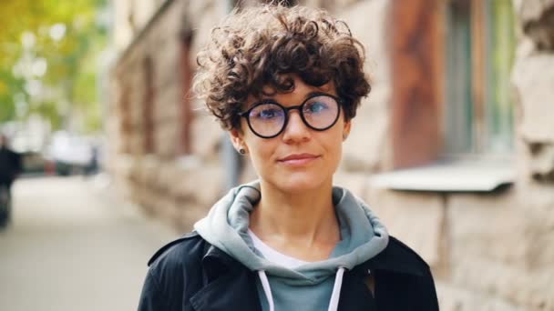 Retrato en cámara lenta de una chica guapa con gafas de pie al aire libre con la cara recta y luego sonriendo y riendo mirando a la cámara. Juventud, emociones y concepto de ciudad . — Vídeos de Stock