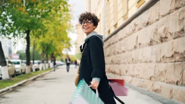 Zeitlupe einer fröhlichen jungen Shopaholic-Frau, die auf der Straße tanzt, helle Einkaufstüten dreht und in die Kamera blickt. Konzept für urbanen Lebensstil und Konsum. — Stockvideo