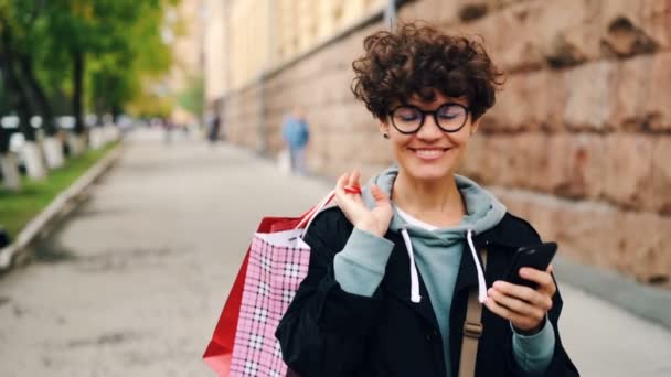 Slow motion of pretty girl with bright paper shopping bags walking in the street and using smartphone then looking around and smiling. Youth lifestyle and city concept. — Stock Video