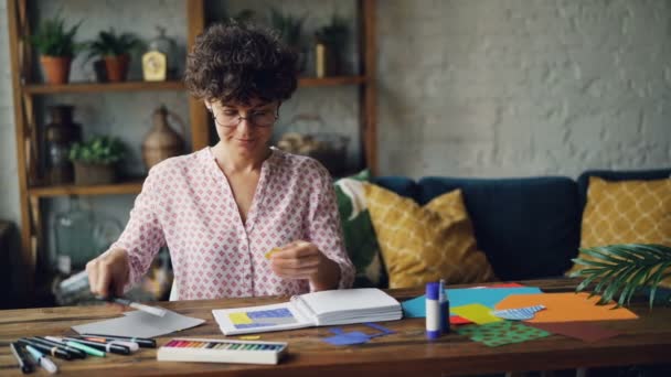 Junge Brünette mit Brille bei der Gestaltung von Illustrationen, die Spritzpapier schneiden und dann mit Klebstoff und Schere Figuren in Notizbücher kleben. Künstler und Kreativitätskonzept. — Stockvideo